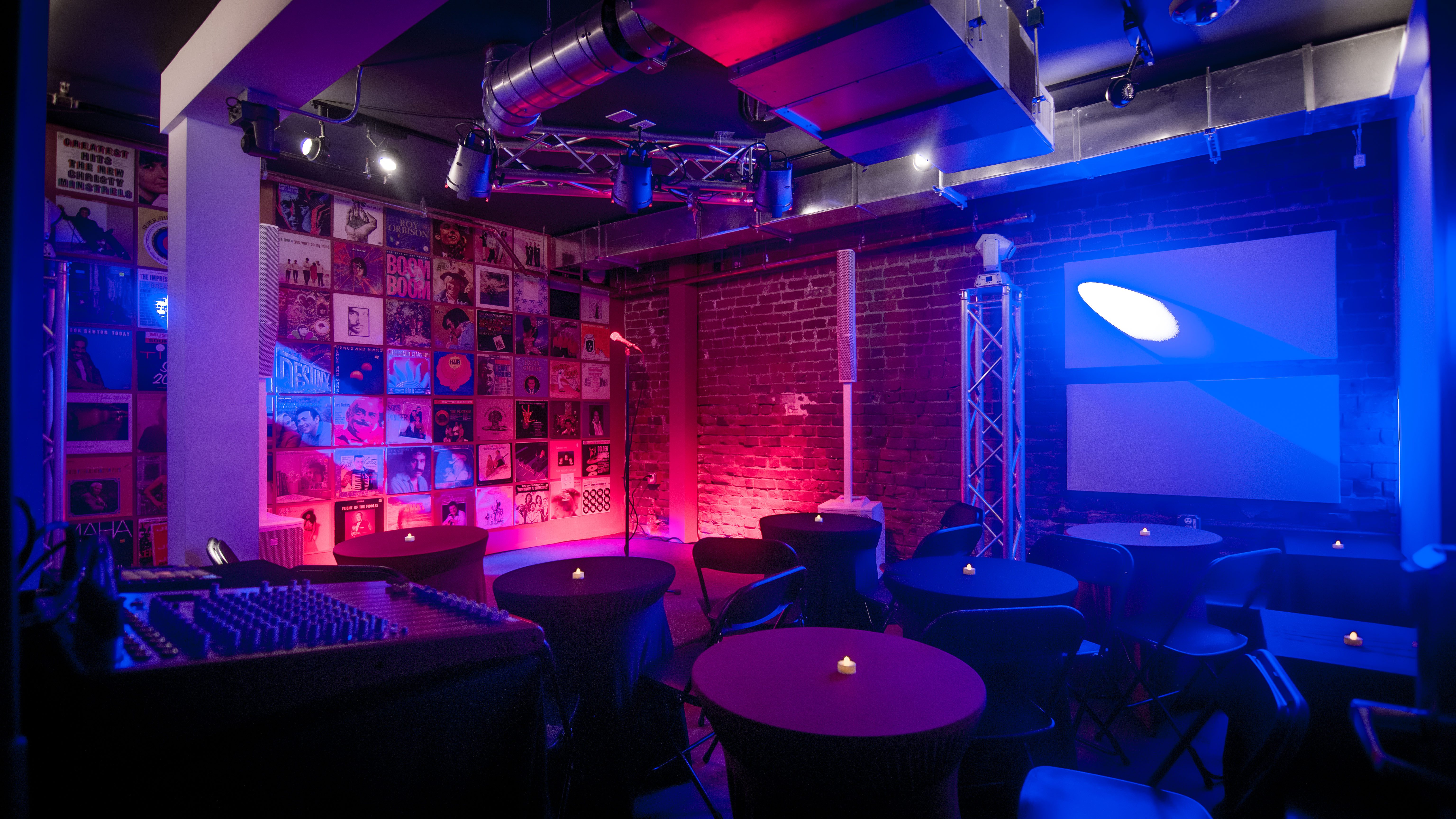 A dimly lit lounge with round tables and chairs, featuring a soundboard and posters on the walls.