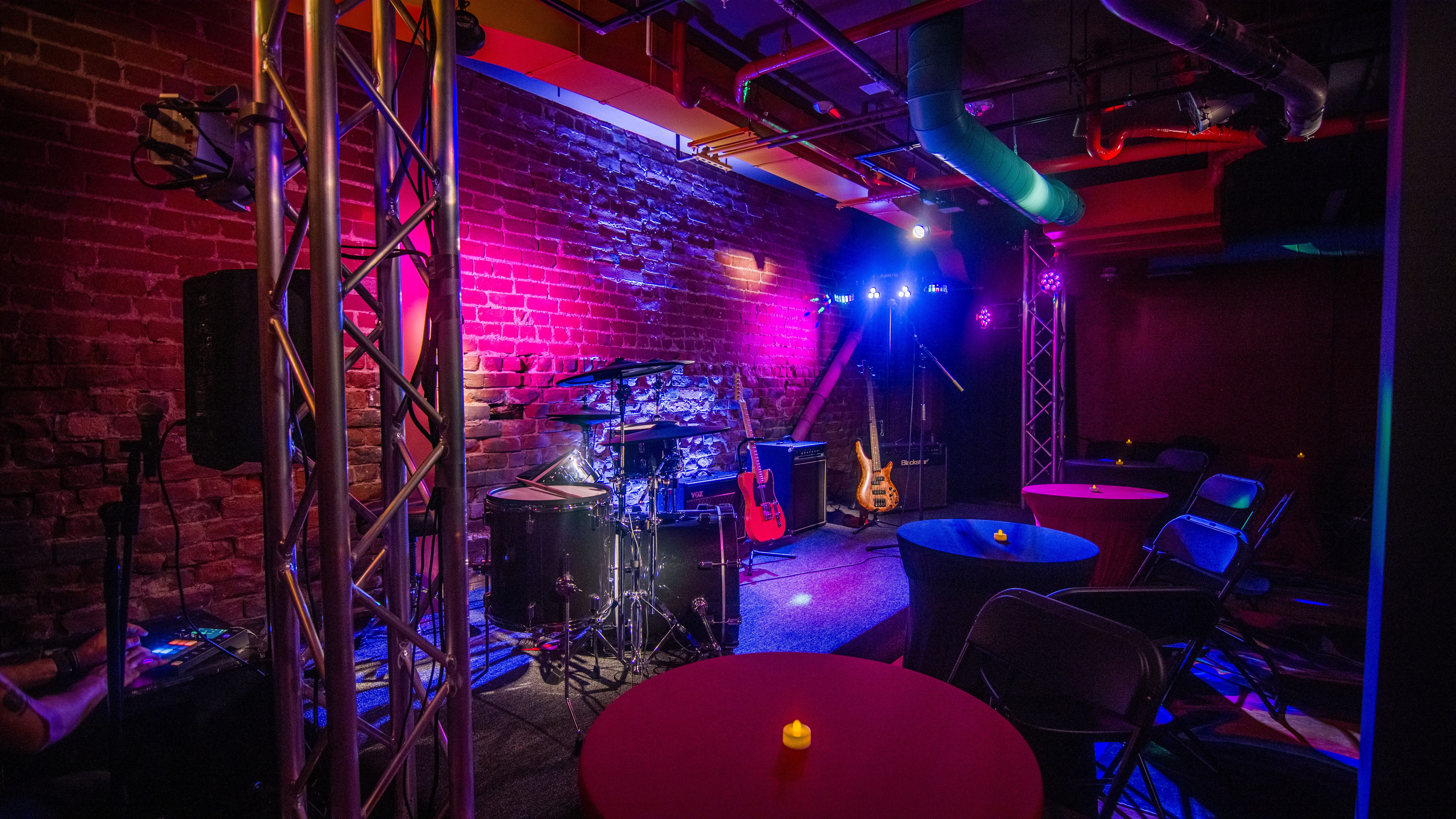 A dimly lit room with a stage, musical instruments, tables, and chairs.
