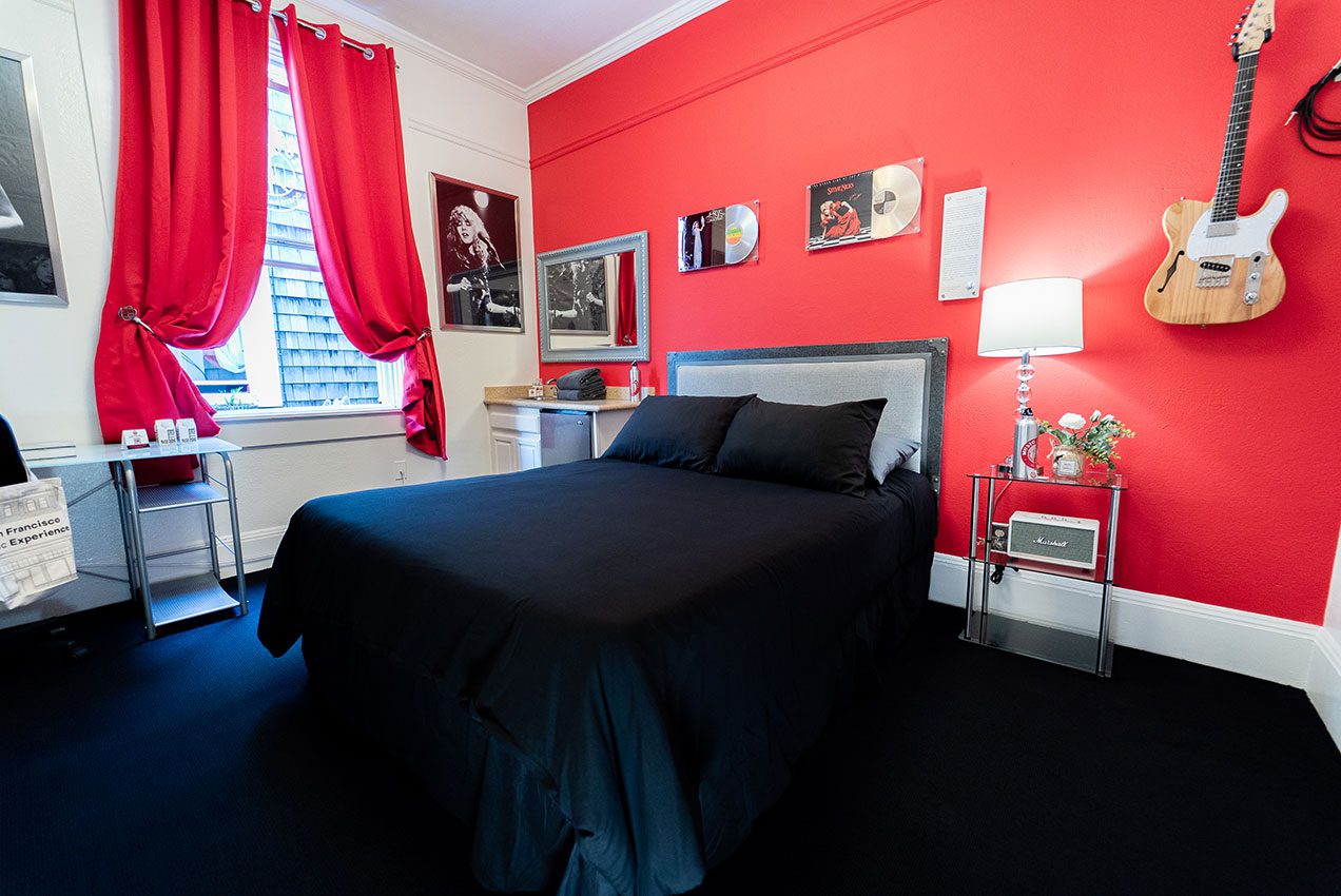 A wide view of a hotel room with a red accent wall, a bed, and a guitar hanging on the wall.